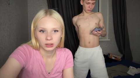 Media: Video of a blonde woman in a pink top and a shirtless man in white pants in a bedroom with gray walls and a window.