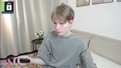 Media: Video of a young man with light skin and short brown hair, wearing a grey t-shirt, playing a video game on a black controller. He sits on a beige sofa in a room with light-colored curtains and a small side table with flowers.