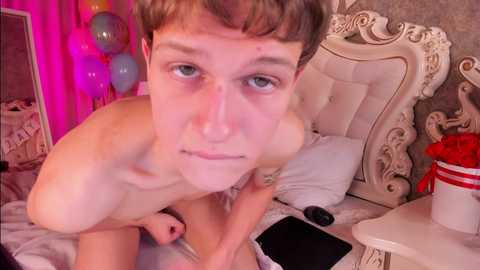 Media: Video of a young, fair-skinned, nude man with short brown hair, kneeling on a bed, with a luxurious, ornate headboard. Background includes balloons, a red gift box, and a mirror.