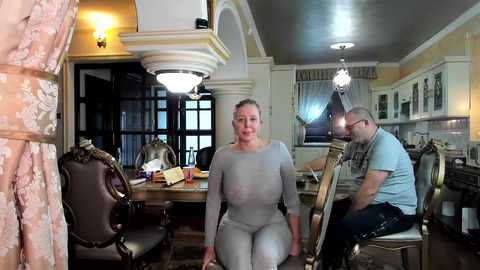 Media: Video of a middle-aged woman with a large bust in a tight gray sweater and high-waisted pants seated at a dining table in a lavish, ornate dining room.