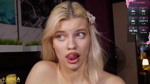 Media: Video of a young blonde woman with fair skin, sticking out her tongue, in a modern room with a digital screen displaying social media metrics.