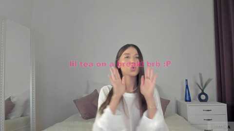 Media: A video of a woman with long brown hair, wearing a white top, standing in a minimalist bedroom with white walls, a white dresser, and a bed with pink pillows.