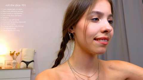 Media: Video of a smiling, light-skinned woman with brown hair in a braid, wearing a strapless top and gold hoop earrings. Background shows a gray curtain and a white dresser with a lamp and decorative items. Text on the left reads \"roll the dip 100.\