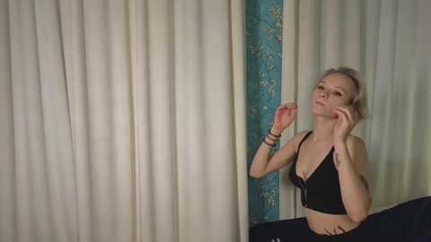 Media: Video of a slim, blonde Caucasian woman with fair skin in a black bra, standing against beige curtains with a teal floral pattern. She has her hand on her cheek.