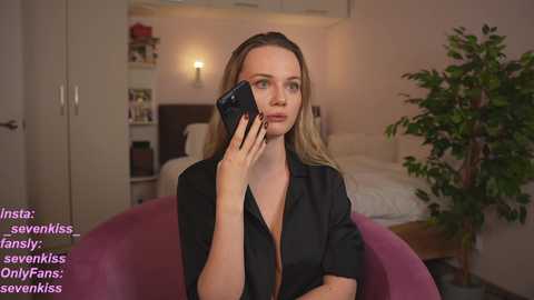 Media: Video of a blonde woman in a black shirt, talking on a phone in a cozy bedroom with a bed, green plant, and pink chair.