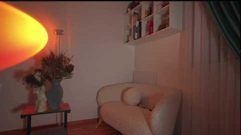 Media: Video of a cozy, dimly lit room with a white curved armchair, a tall vase of flowers, and a shelf filled with books. A large, bright orange sun-shaped lamp casts a warm glow on the left wall.