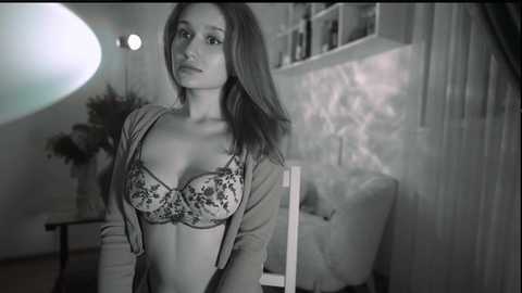 Media: Black-and-white video of a young, slender woman with long hair, wearing a floral-patterned bra and open cardigan, standing in a dimly lit room with a bookshelf and potted plant in the background.