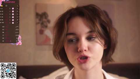 Media: Video of a young woman with short, tousled brown hair, wearing a white blouse, looking surprised. Background shows a blurred, beige wall with a painting. QR code in the bottom left corner.