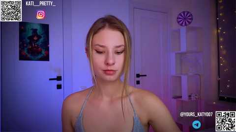 Media: Video of a young woman with fair skin, straight blonde hair, and light makeup, wearing a gray spaghetti-strap top, in a dimly lit room with purple lights, framed art, and a dartboard.