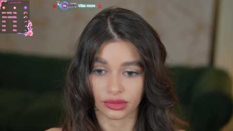 Media: Video of a young woman with long, wavy dark hair, wearing minimal makeup and a pink top, set against a blurred green couch.