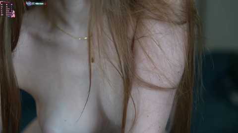 Media: Video of a fair-skinned, topless woman with long, straight, reddish-brown hair, wearing a gold necklace, taken from the neck down, in a dimly lit room with a blurred background.