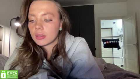 Media: A video of a young woman with long, wavy brown hair, fair skin, and a neutral expression, wearing a grey robe, lying on a bed in a dimly lit bedroom with white walls and a door open to a closet.