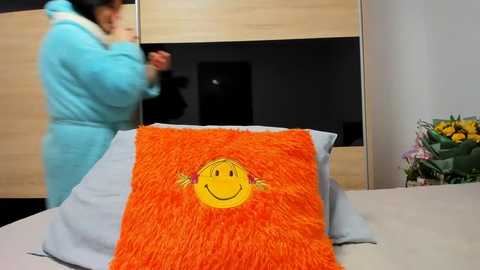 Media: Video of a woman in a blue robe adjusting a fuzzy, orange blanket with a smiley face embroidered on it, in a modern bedroom with light wooden furniture and floral decorations.