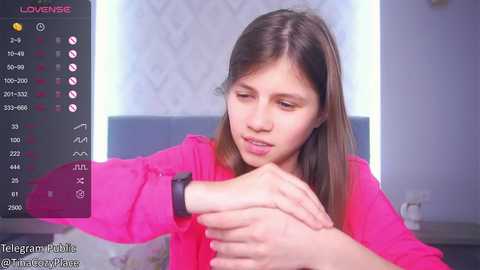 Media: Video of a young woman with straight brown hair, wearing a bright pink top, looking at her wristwatch, in a modern room with white walls and a grey bed.