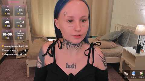 Media: Video of a young woman with blue hair and multiple tattoos, including \"Loti\" on her chest, sitting in a beige room with a bed and a lamp.