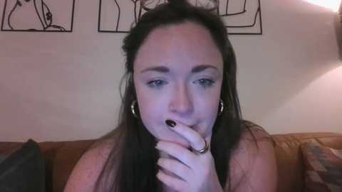 Media: A video of a young woman with long, dark hair, fair skin, and light makeup, biting her fingernail. She wears hoop earrings and is seated on a brown leather couch in a dimly lit room.