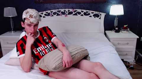 Media: Video of a young man with short, bleached hair wearing a red and black Inter Milan jersey, beige shorts, and a backward cap, lying on a white bed in a dark bedroom with a white headboard, lamps, and a cluttered nightstand.