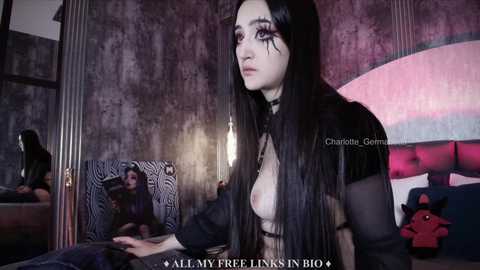 Media: Video of a young woman with long black hair, wearing a sheer black top revealing her small breasts, in a dimly lit, gothic-themed bedroom with zebra-patterned bedding and a large, red plush toy.