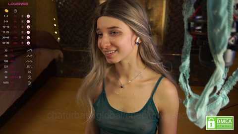 Media: Video of a smiling young woman with light skin and long blonde hair, wearing a sheer teal tank top, sitting on a wooden floor in a dimly lit room.