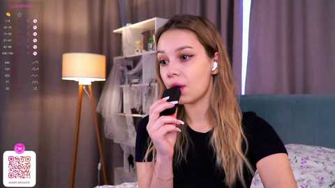 Media: Video of a young woman with long blonde hair, wearing a black top, applying lipstick in a bedroom with a bed, lamp, and window curtains.