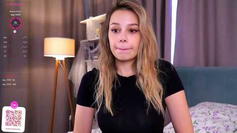 Media: Video of a young woman with long blonde hair, wearing a black top, sitting on a bed with floral sheets. Background includes a lamp and gray curtains.