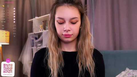 Media: Video of a young girl with long, wavy blonde hair and light skin, wearing a black top, applying makeup in a well-lit bedroom with neutral-toned curtains and a blue couch.