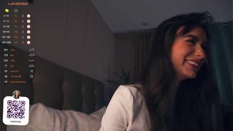 Media: Video of a smiling woman with long dark hair, wearing a white top, in a dimly lit bedroom with a bed and curtains in the background.