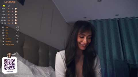 Media: Video of a smiling woman with long, dark hair, wearing a white top, sitting on a bed with teal curtains in the background.