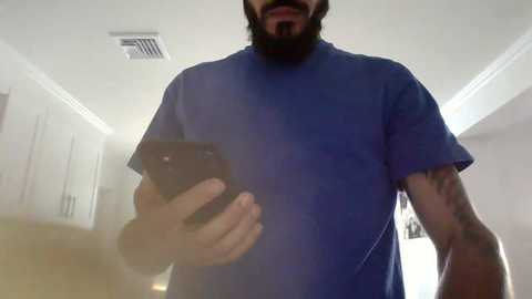 Media: A video of a man with a beard, wearing a blue t-shirt, taking a selfie with a black smartphone, displaying a tattoo on his right arm. The background features a white ceiling and white cabinets.