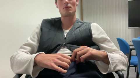 Media: Video of a middle-aged Caucasian man with short hair, wearing a white long-sleeved shirt and dark gray vest, sitting in a chair. He appears to be adjusting his pants. Background includes a white wall and a blue chair.