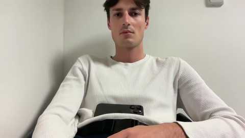 Media: A video of a young man with short dark hair, wearing a white ribbed sweater, seated in a corner of a plain, white-walled room. He holds a black smartphone.