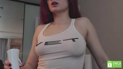 Media: A video of a fair-skinned woman with long, red hair, wearing a white tank top that reveals her large breasts, standing in a modern bedroom.