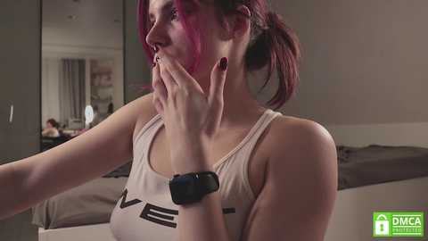 Media: Video of a woman with pink hair, wearing a white tank top and black smartwatch, covering her mouth with her hand, standing in a modern bedroom.