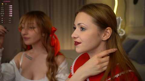 Media: Video of two young women in a dimly lit room. One has long, straight, reddish-brown hair and wears a red dress, while the other has wavy auburn hair, a white top, and a red bow. They're engaged in conversation.