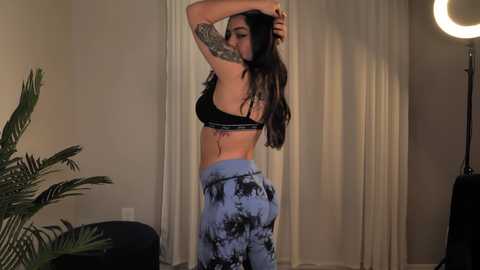 Media: Video of a curvaceous woman with long, dark hair, wearing a black bra and blue tie-dye pants, posing in a dimly lit room with beige curtains and a potted plant.