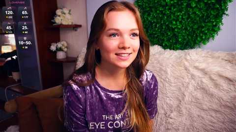 Media: Video of a young, fair-skinned, red-haired girl with blue eyes, smiling, wearing a velvet purple shirt, in a cozy room with a white fur blanket, green wall art, and a TV screen displaying a weather app.