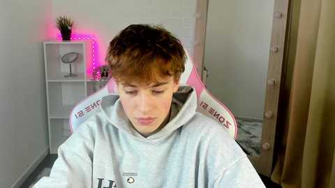 Media: Video of a young boy with curly brown hair, wearing a light gray hoodie, sitting in a gaming chair, in a modern, brightly lit room with pink LED strip lights and a white shelf.