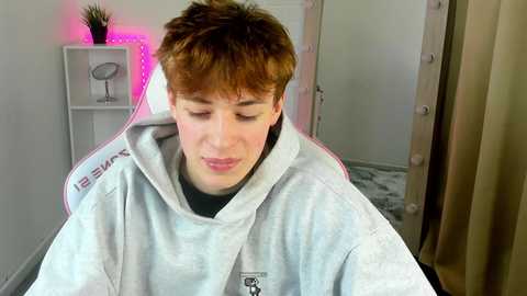 Media: Video of a young man with short, tousled brown hair, wearing a light grey bathrobe, standing in a modern bathroom with white walls and a mirror, illuminated by pink LED lights.