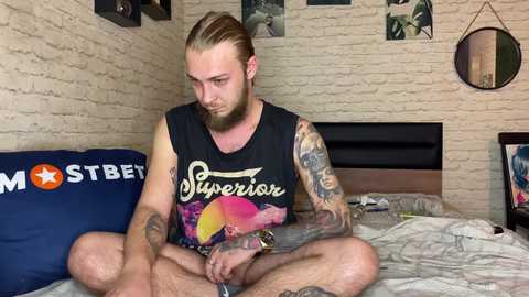 Media: Video of a tattooed man with long hair, sitting cross-legged on a bed with a black tank top, in a cozy, dimly-lit room with exposed brick walls and a Star Trek-themed pillow.