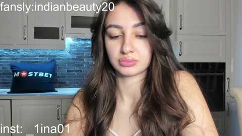 Media: Video of a young woman with long, wavy brown hair, fair skin, and a soft expression. She's wearing a white tank top. Background shows a modern kitchen with grey cabinets, a brick backsplash, and a blue cushion.