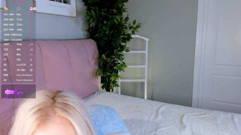 Media: A video of a bedroom with a light-skinned woman with blonde hair partially visible, lying on a bed with a white and light blue bedspread. The room has a white door and a green leafy plant.