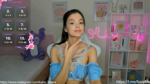 Media: A young woman with long black hair, wearing a blue off-shoulder checkered top, sits on a white chair in a cozy room with pink neon lights, digital frames, and a purple plush toy.