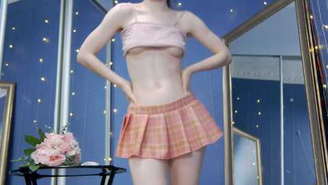 Media: Video of a slender, pale-skinned woman wearing a pink plaid skirt and a cropped, pink spaghetti-strap top, standing with hands on hips. Background includes a mirror and a flower arrangement on a table.