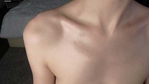 Media: Video of a light-skinned, bare-chested person from the shoulders down, showing a prominent shoulder blade. Background includes a gray carpet and a blurred bed with white sheets. The image has a soft, natural lighting.