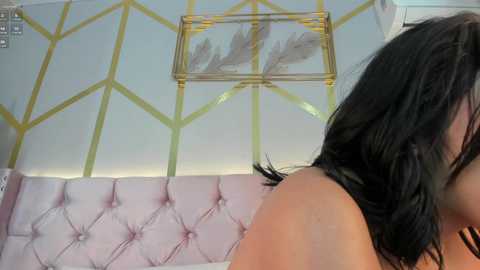 Media: Video of a woman with long black hair, seen from the side, in a room with a pink, tufted sofa, a yellow geometric wall, and a framed feather artwork.