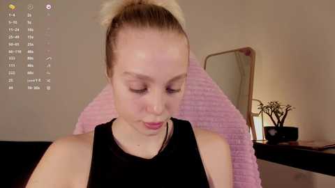 Media: Video of a fair-skinned woman with light brown hair in a high ponytail, wearing a black tank top, seated in a pink cushioned chair. Background includes a desk with a plant and a mirror.