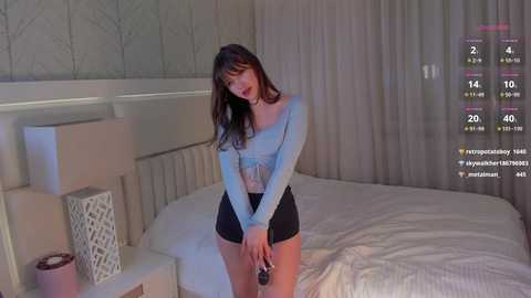 Media: Video of a slender, fair-skinned woman with long brown hair, wearing a light blue, long-sleeved top and black shorts, standing in a modern, beige-toned bedroom with white curtains. She holds a phone, and a virtual camera overlay shows her face and body.