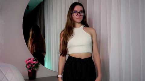 Media: Video of a young woman with long brown hair, wearing glasses, a white crop top, and black pinstripe pants, standing in front of a circular mirror with pink flowers in a vase.