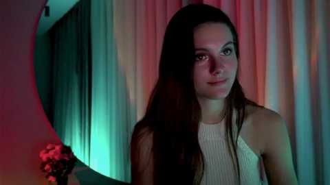 Media: Video of a young woman with long, dark hair, wearing a white sleeveless top, standing against a backdrop of colorful, draped curtains.