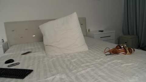 Media: Video of a minimalist bedroom with a white bed, a toppled pillow, a keyboard, and a pair of brown sandals on the floor, against a plain white wall.